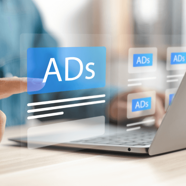 A hand points at a floating digital interface displaying 'ADs,' in front of a laptop on a wooden desk.