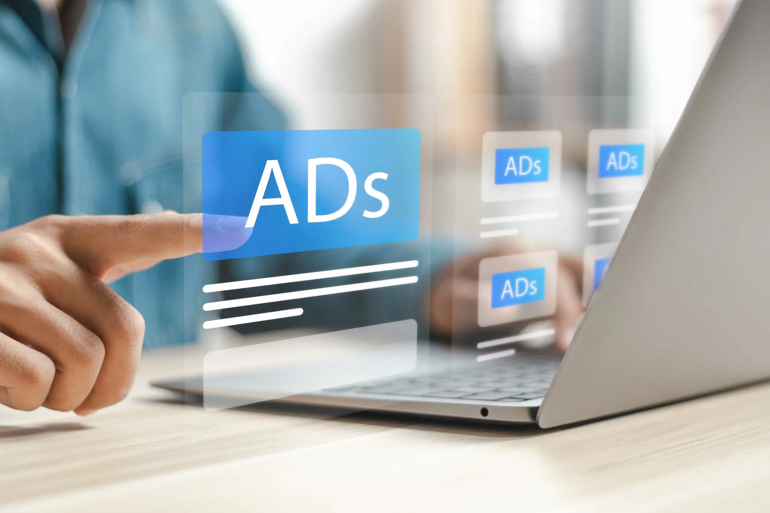 A hand points at a floating digital interface displaying 'ADs,' in front of a laptop on a wooden desk.